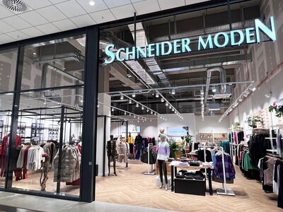 Innenansicht eines Modegeschäfts mit Kleiderauslagen und der Aufschrift „Schneider Moden“.