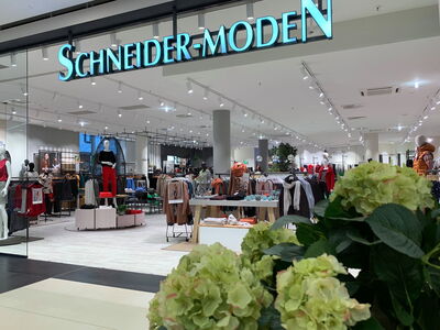 Innenansicht eines Modegeschäfts mit dem Schriftzug „Schneider-Moden“ und ausgestelltem Kleidungsstücken.